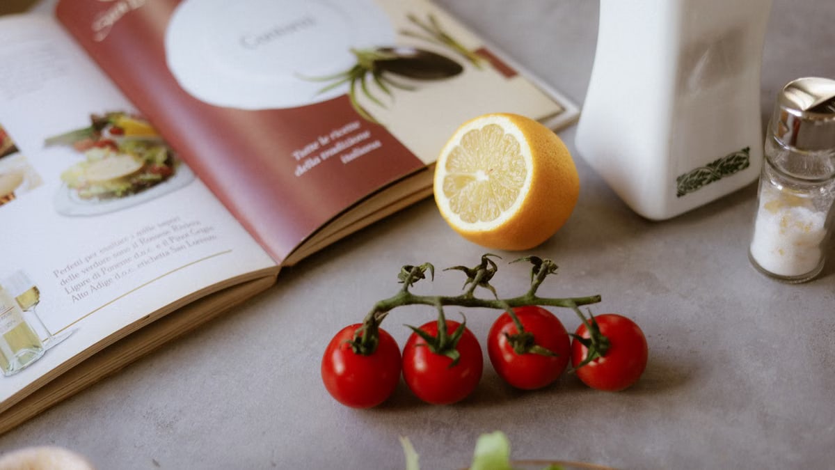 Flat lay of cooking ingredients including tomatoes and a lemon with a cookbook.